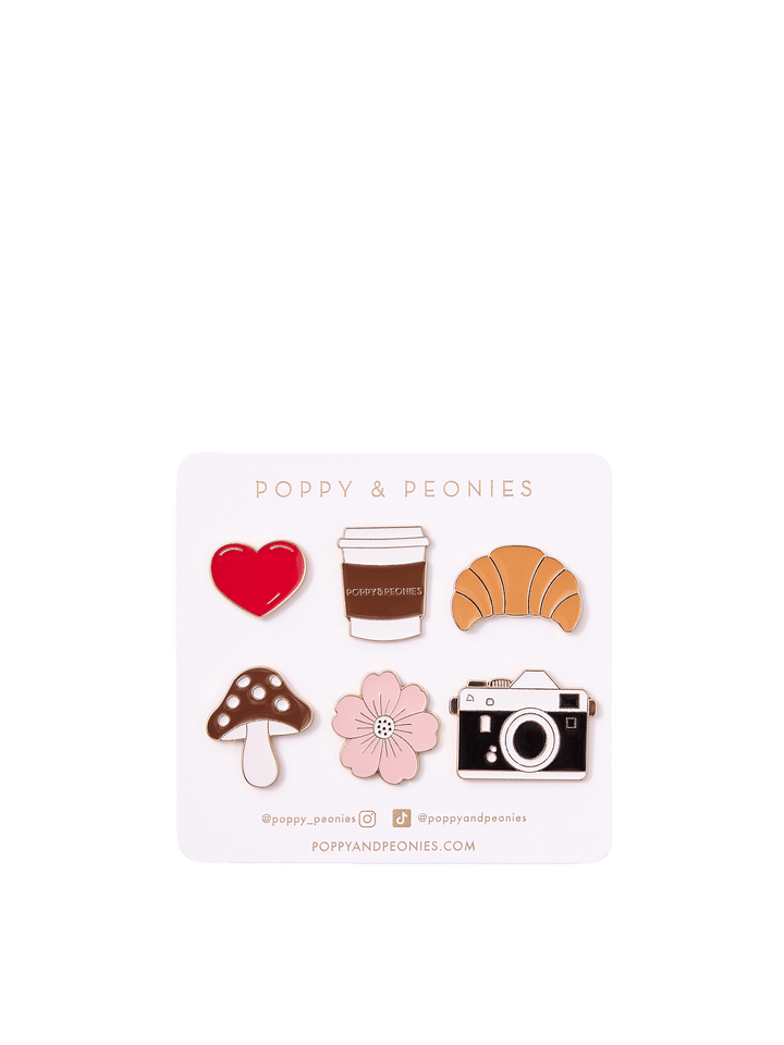 Poppy & Peonies Enamel Pin Set featuring six playful designs: a red heart, takeaway coffee cup, croissant, brown mushroom, pink flower, and vintage camera, displayed on a branded backing card.