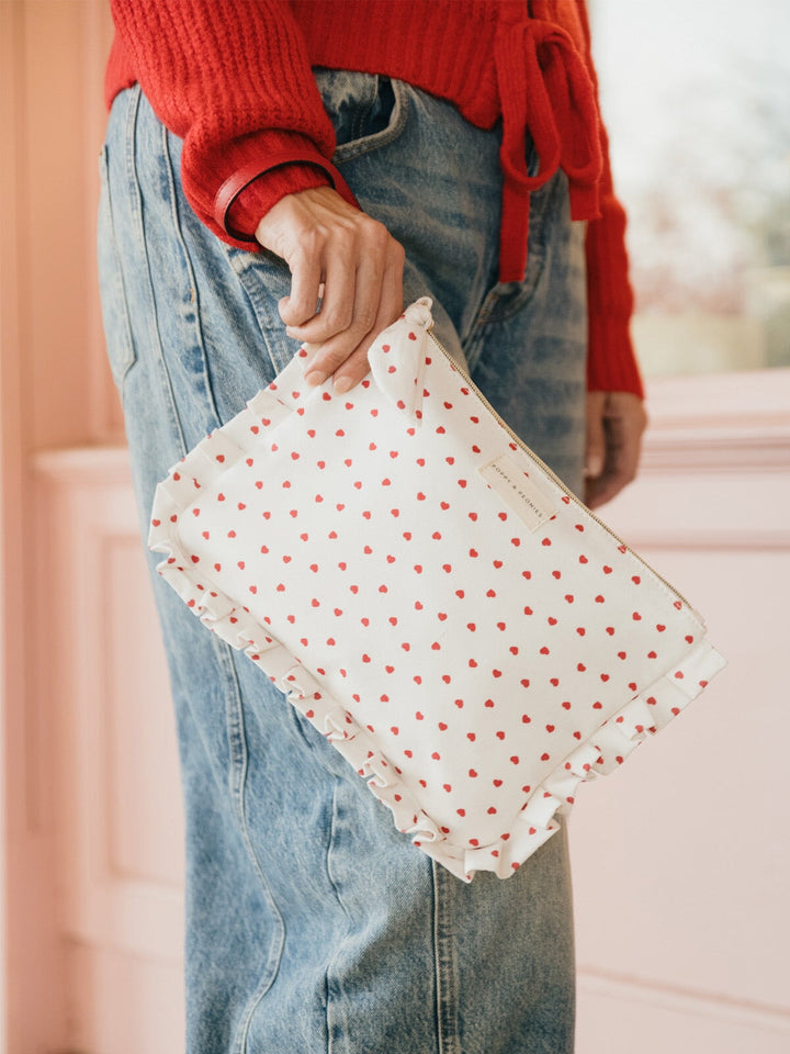 Person holding a white clutch with red heart pattern and red wrist strap, wearing a red sweater and blue jeans.