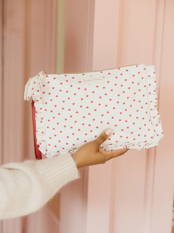 Hand holding a white clutch with red heart pattern and red wrist strap against a pink curtain background