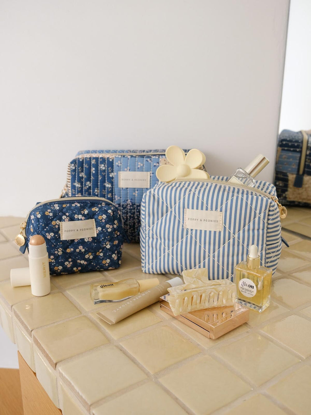 Product-focused lifestyle shot of the Cosmetic Trio in Navy Patchwork, featuring all three coordinating cosmetic cases in blue floral, striped, and patchwork prints styled alongside beauty essentials.