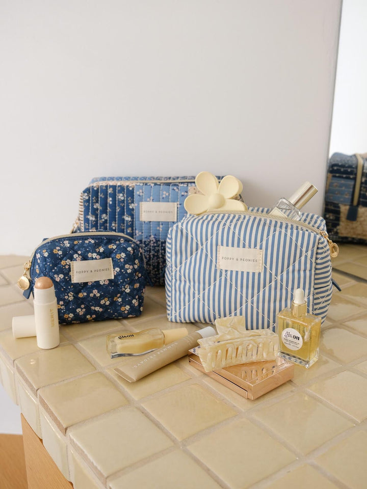 Product-focused lifestyle shot of the Cosmetic Trio in Navy Patchwork, featuring all three coordinating cosmetic cases in blue floral, striped, and patchwork prints styled alongside beauty essentials.