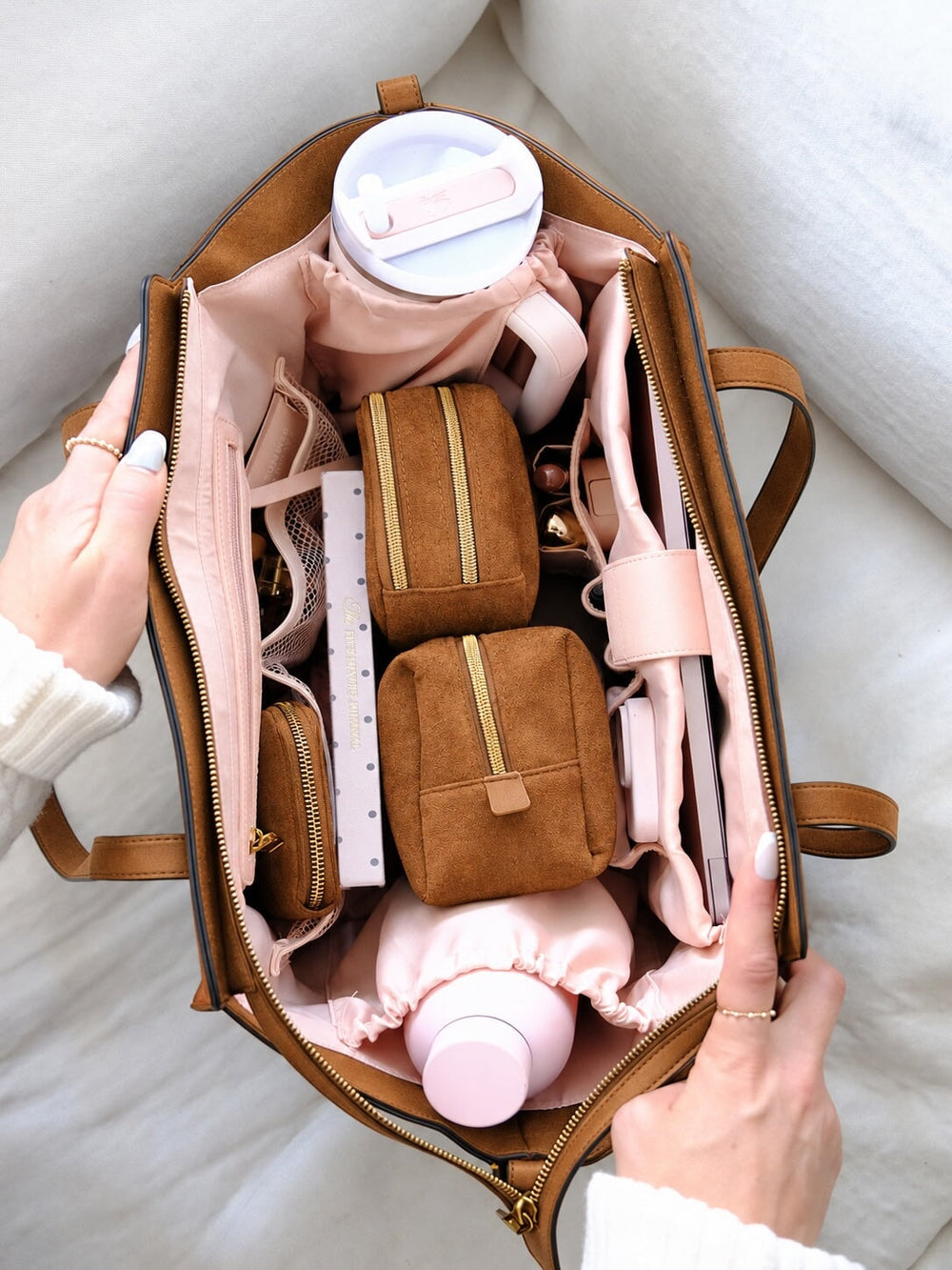 An open brown suede tote bag filled with matching pink and brown travel accessories, including pouches, a water bottle, and a phone strap, held open by two hands over a soft white fabric background.