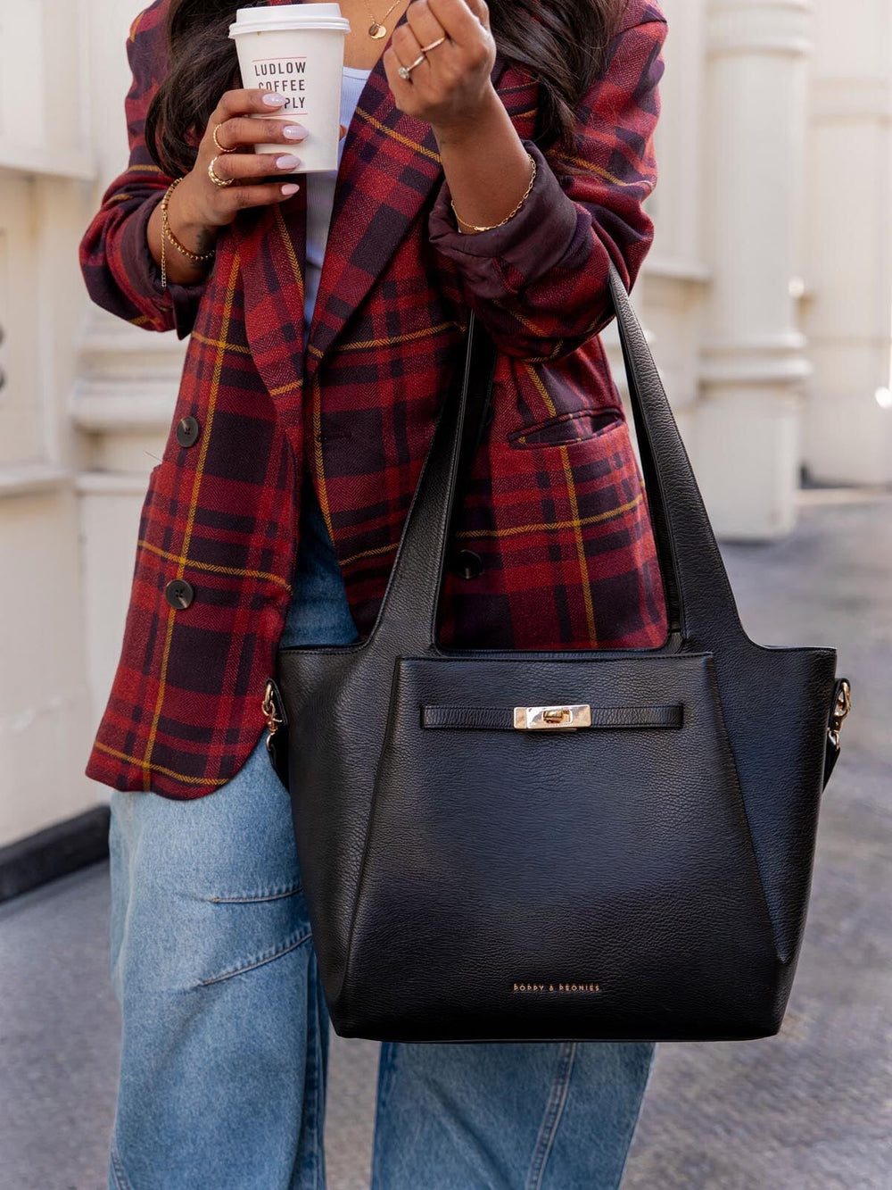 Woman wearing a plaid jacket and holding a black vegan leather tote bag with gold hardware in an urban setting