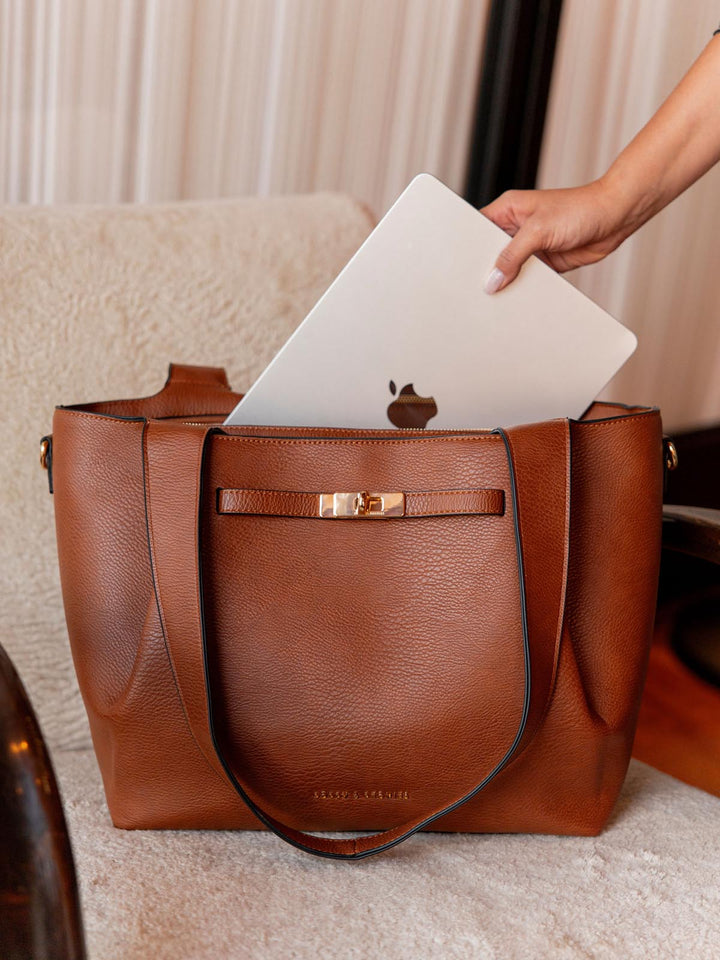 Brown vegan leather handbag with a laptop being placed into it on a light-colored surface.