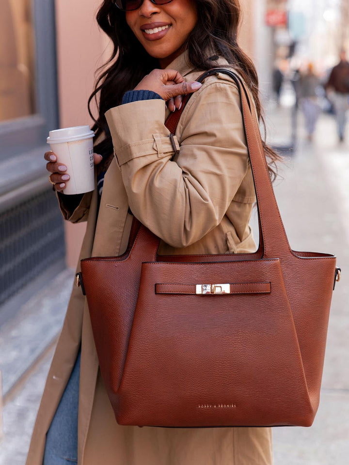 Woman holding a brown vegan leather tote bag with gold hardware and a coffee cup on a city street.