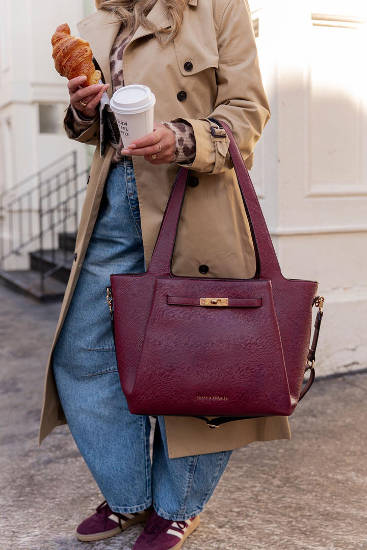 Person holding a coffee cup and a croissant, wearing a beige coat and blue jeans, with a berry coloured vegan leather tote bag with gold hardware.