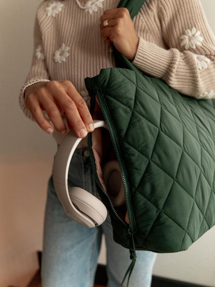 Person holding a green quilted bag and white headphones, wearing a beige sweater with floral patterns.