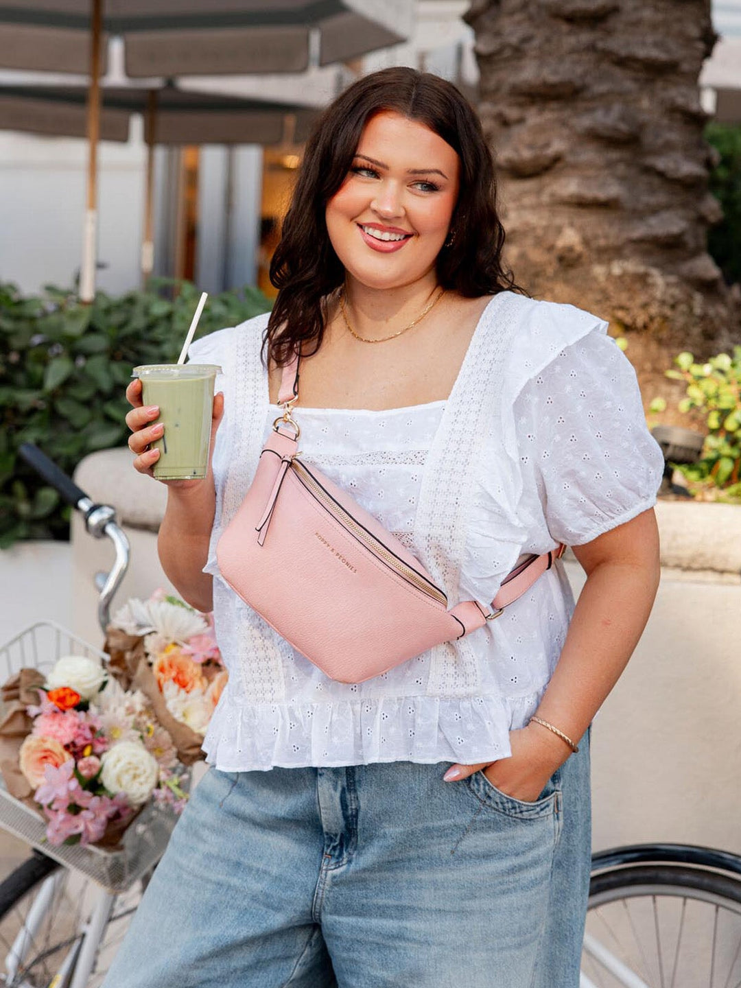 Woman holding a drink and a pink bag, standing outdoors with a bicycle and flowers in the background.