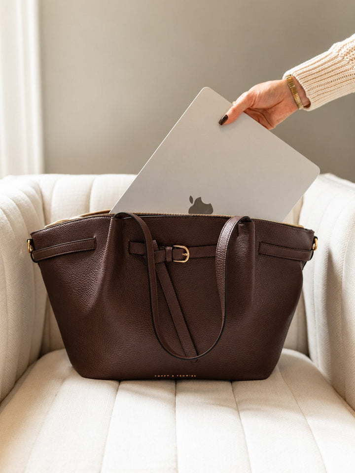 Poppy & Peonies The Knot Tote in Espresso, resting on a cream armchair as a hand places a silver laptop into its spacious main compartment.