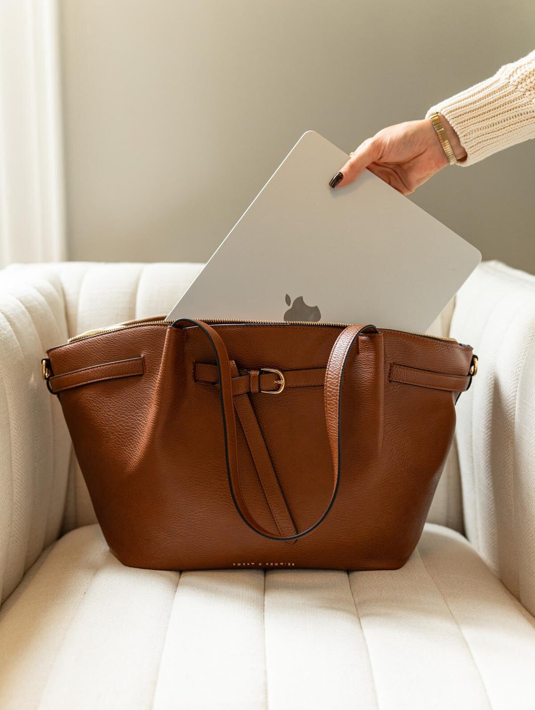 Poppy & Peonies The Knot Tote in Chestnut, resting on a cream armchair as a hand places a silver laptop into the main compartment.