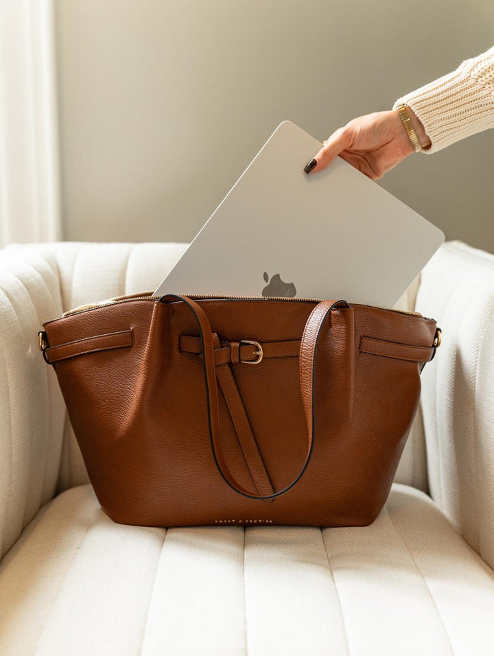 Poppy & Peonies The Knot Tote in Chestnut, resting on a cream armchair as a hand places a silver laptop into the main compartment.