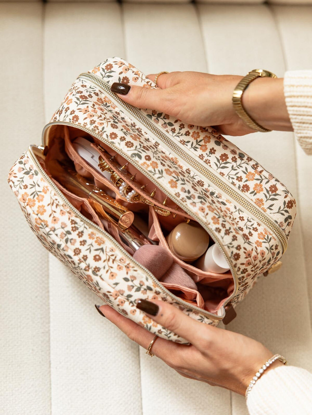 Poppy & Peonies Double Take Cosmetic Case in October Floral, shown open on a cream chair with blush pink lining, multiple compartments, and makeup essentials organized inside.