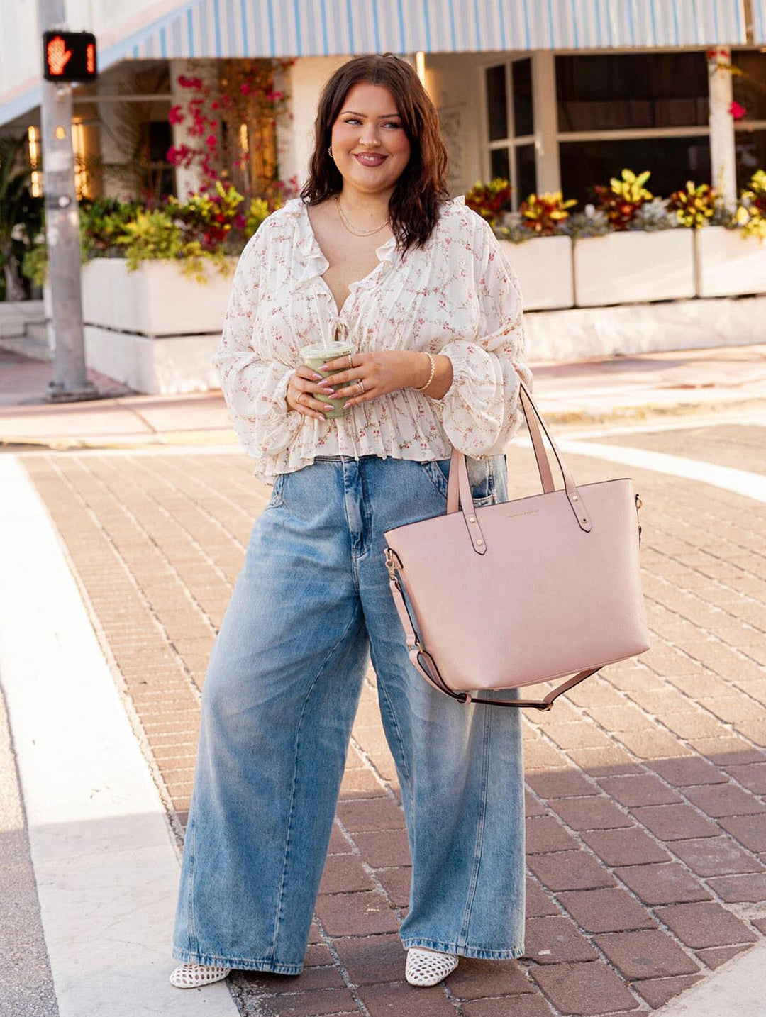 Woman in a white blouse and blue jeans holding a light pink tote bag on a sidewalk.