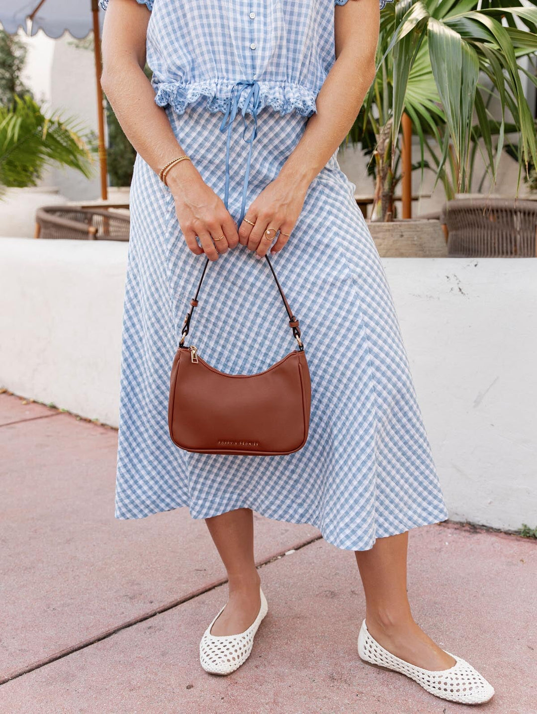 Person wearing a blue checkered dress holding a brown vegan leather handbag on a sidewalk.