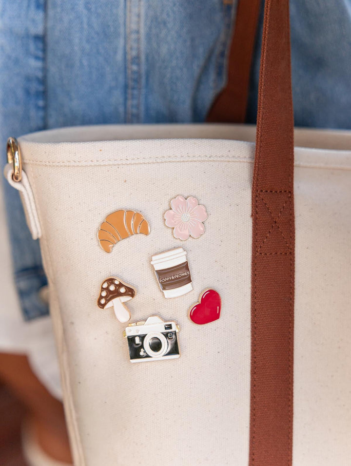 Poppy & Peonies Signature Pins displayed on Signature Tote, featuring a croissant, pink flower, coffee cup, mushroom, camera, and red heart beside a brown shoulder strap.