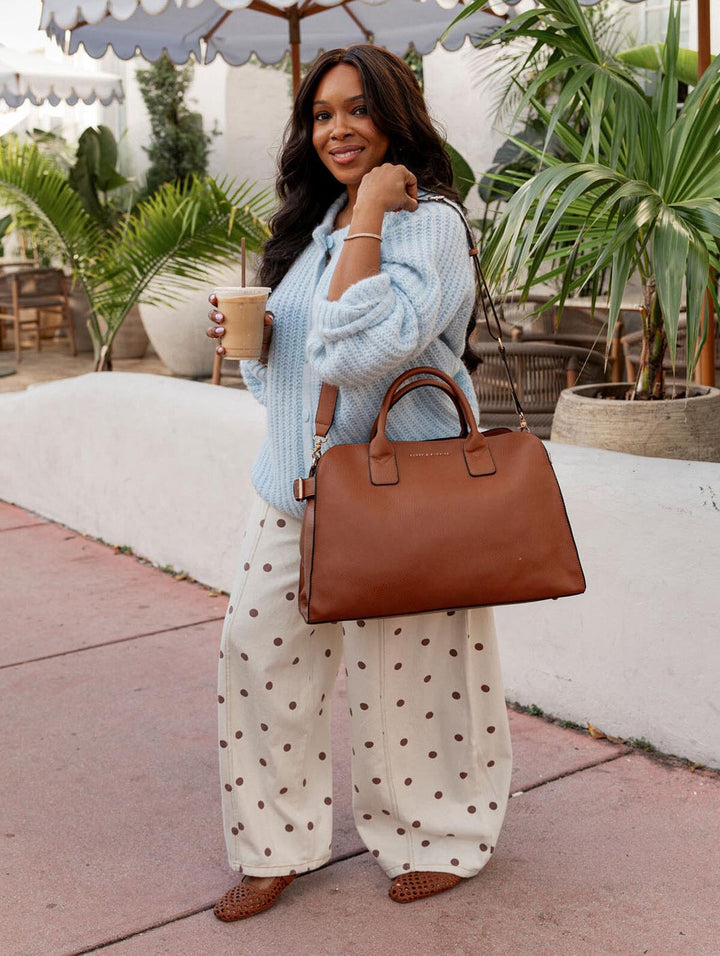 Woman holding a brown handbag and a drink, standing outdoors with plants and tables in the background.