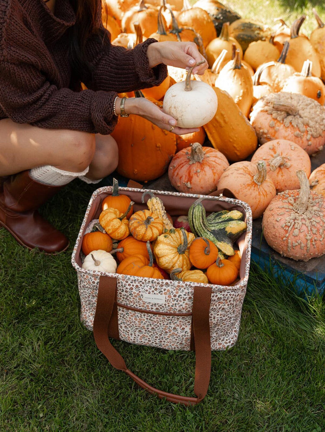 Poppy & Peonies Mini Traveller in October Floral, standing open on the grass at a pumpkin patch with small pumpkins and gourds inside, featuring brown canvas straps and a delicate floral print.