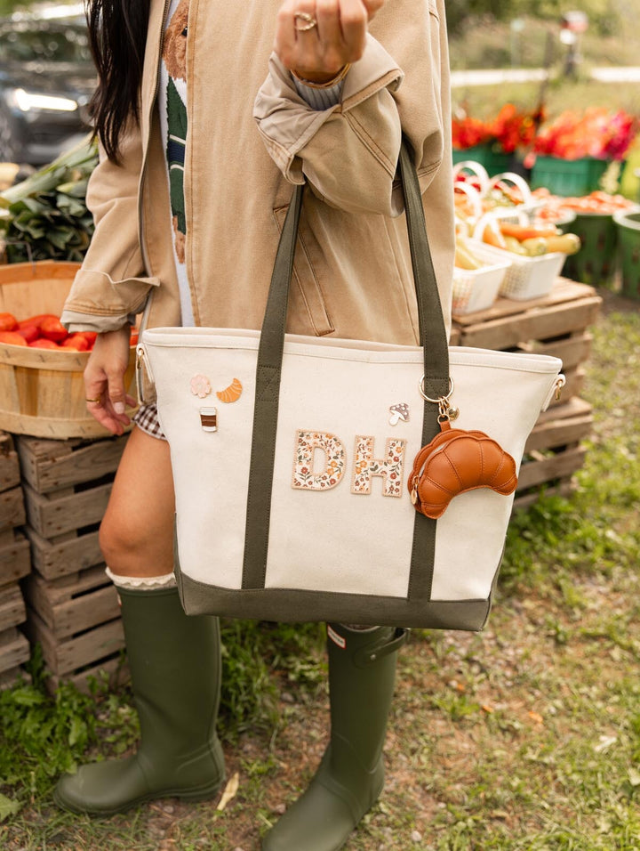 Poppy & Peonies The Signature Tote in Olive carried at an outdoor market, featuring personalized initials and decorative patches on its cream canvas exterior.
