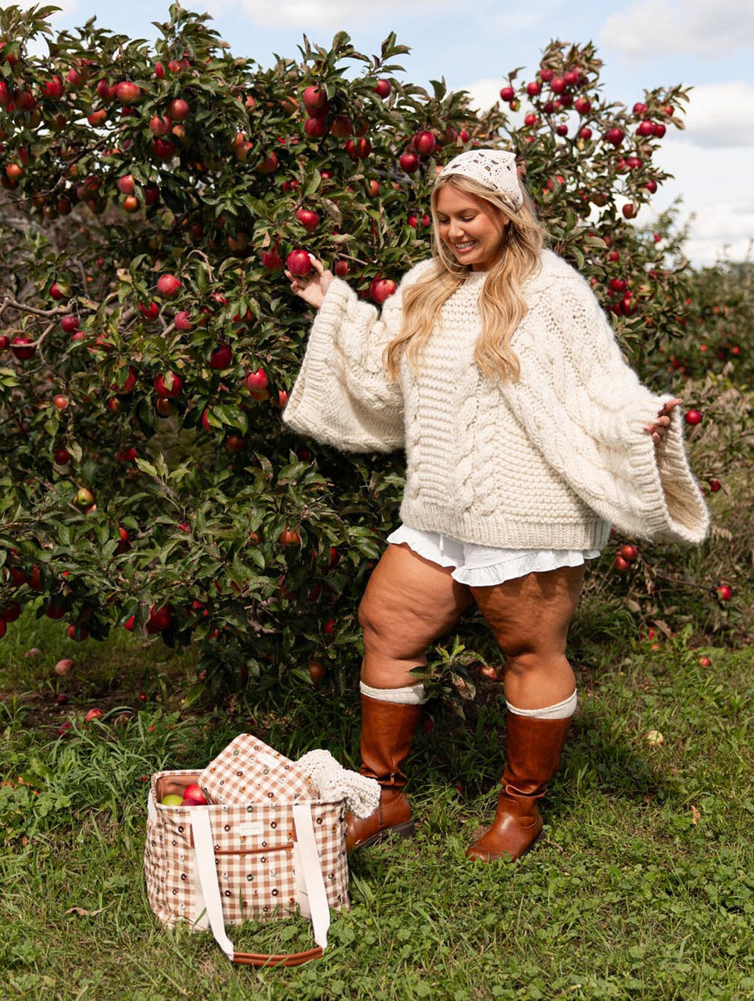 Poppy & Peonies Mini Traveller in October Gingham, carried by a smiling woman in a cream knit sweater and brown boots, featuring its floral gingham pattern and long shoulder straps.