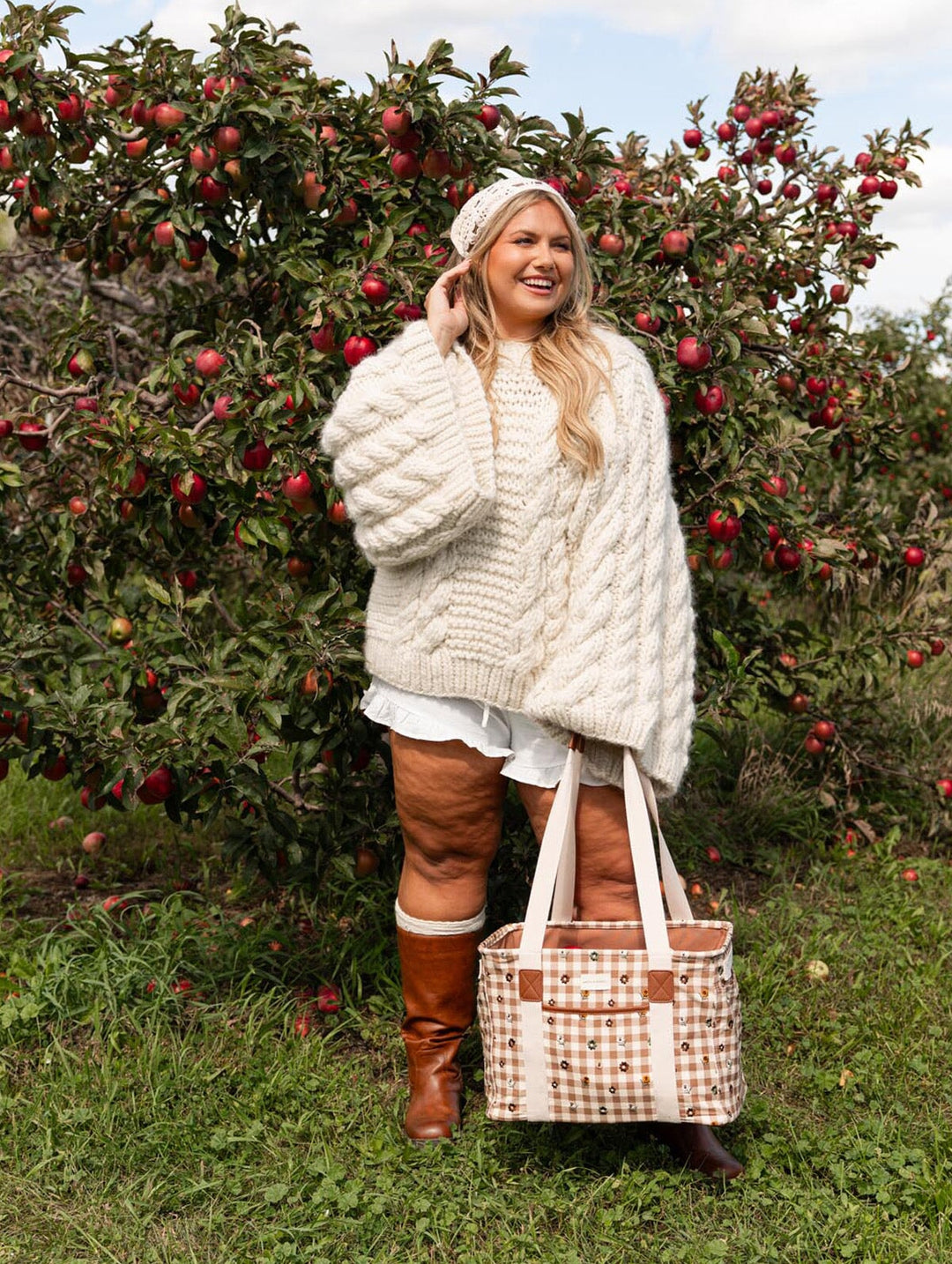 Poppy & Peonies Mini Traveller in October Gingham, placed on the grass with apples peeking from the open top while a woman reaches toward an apple tree.