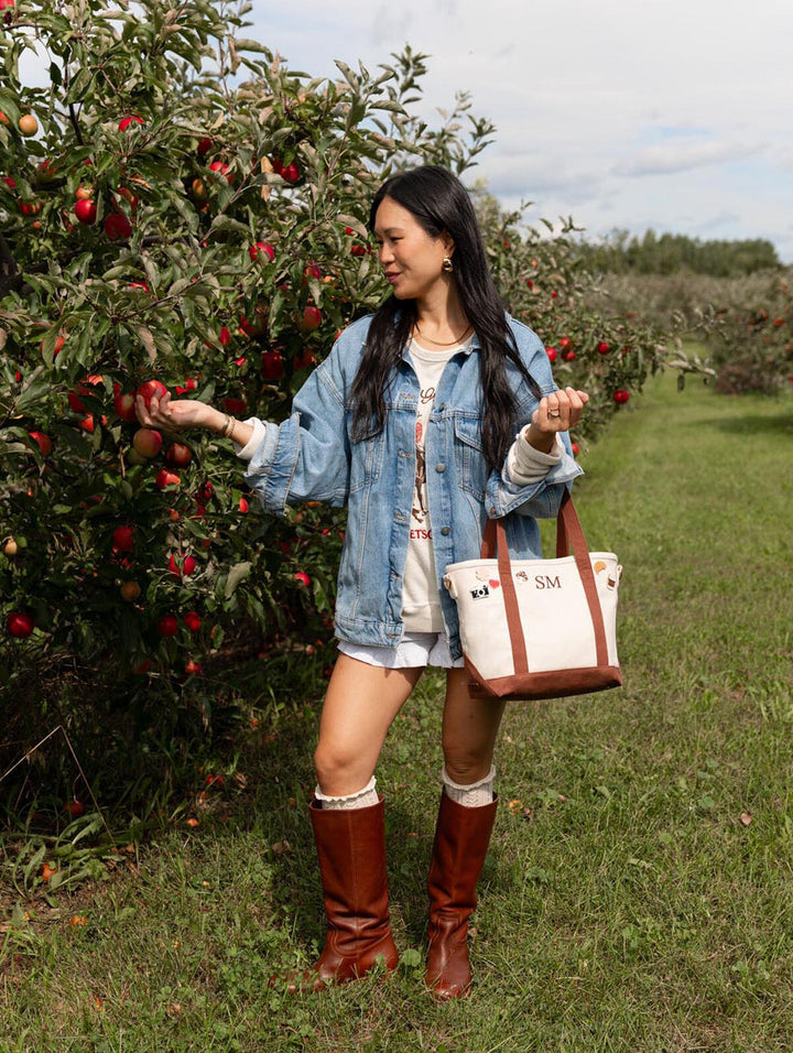 Woman looking at apples on a tree while carrying Poppy & Peonies The Small Signature Tote in Chestnut by its handles.