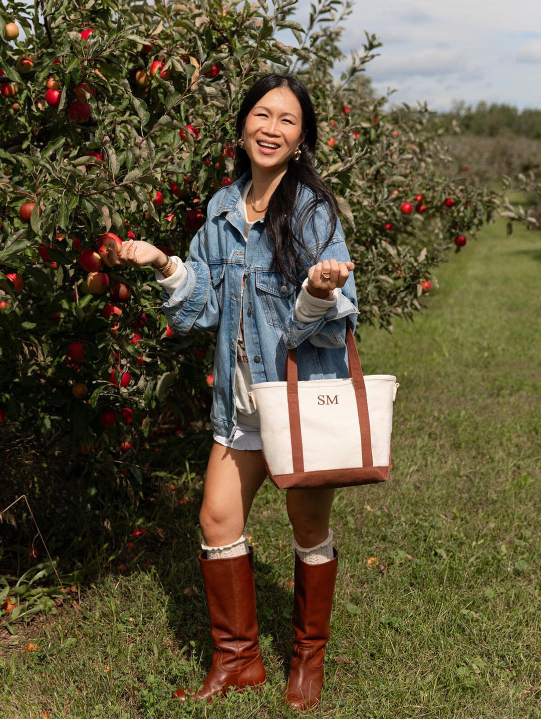 Woman smiling in an apple orchard while holding Poppy & Peonies The Small Signature Tote in Chestnut, styled with a denim jacket and tall brown boots.