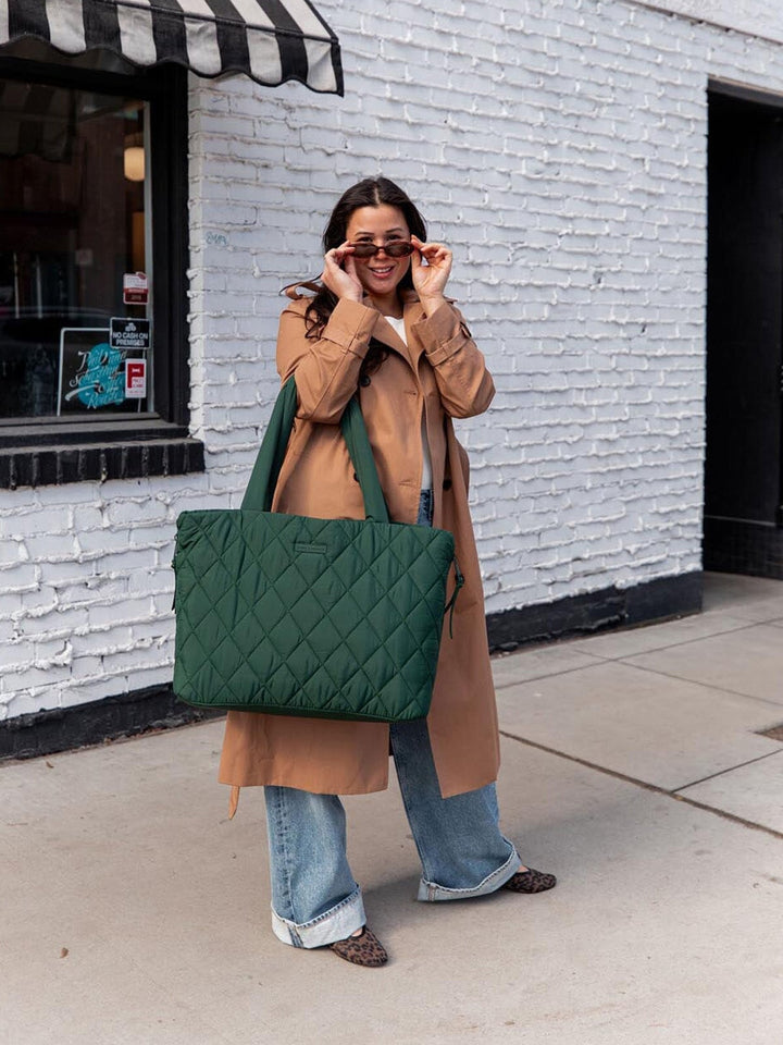 Woman holding a green quilted tote bag in front of a white brick wall.