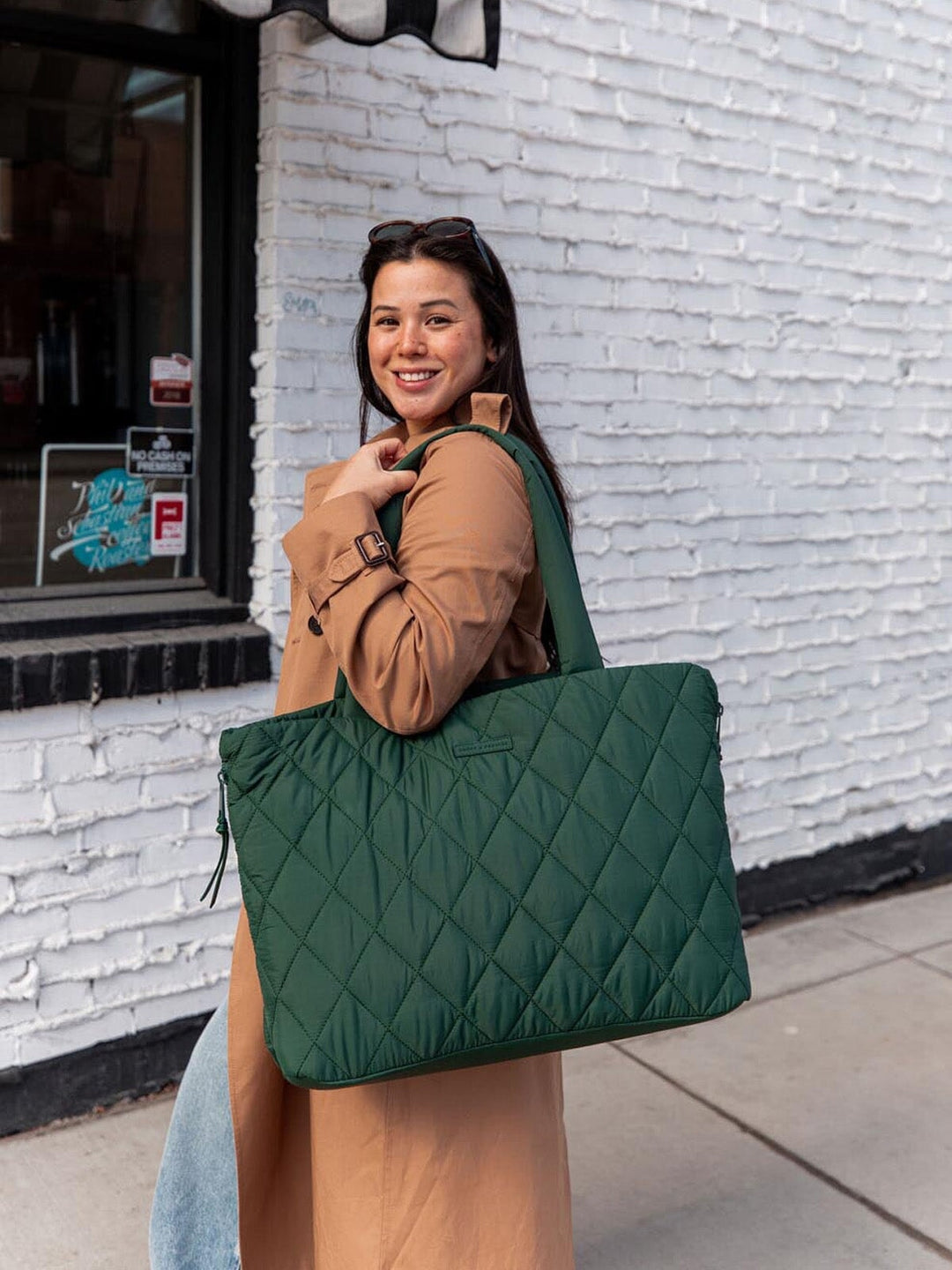 Woman holding a green quilted tote bag against a white brick wall.