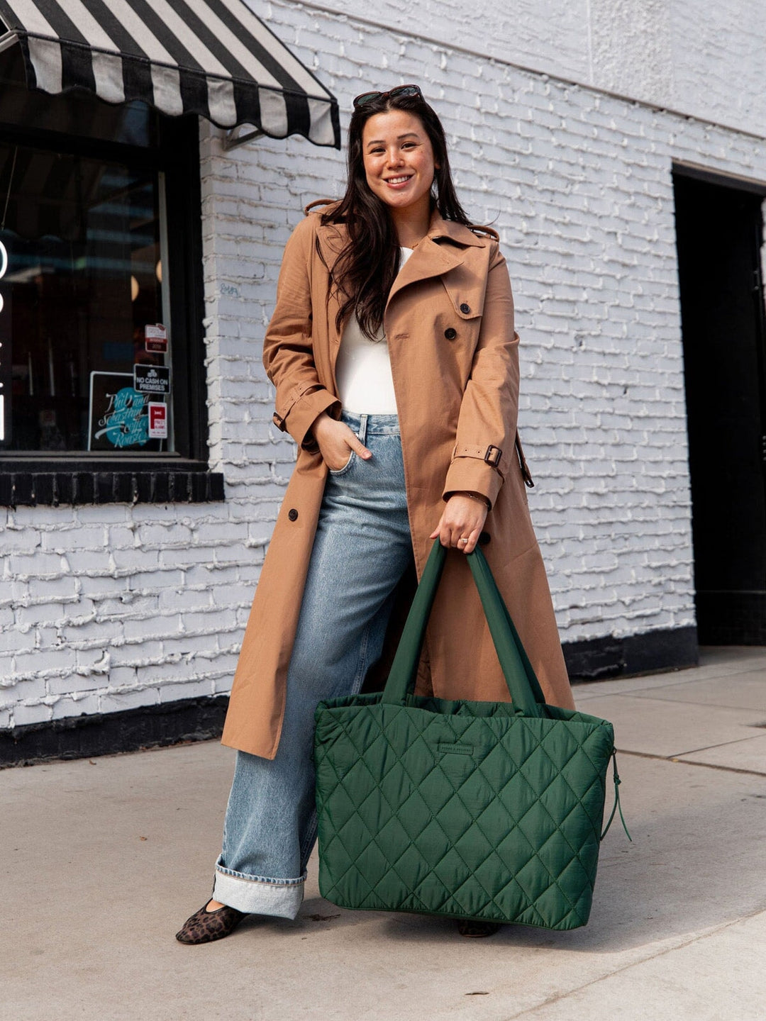 Woman in a tan coat holding a green quilted tote bag against a white brick wall.