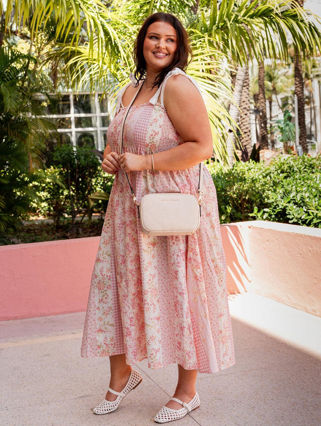 Woman in a pink floral dress holding a cream crossbody bag outdoors with greenery in the background