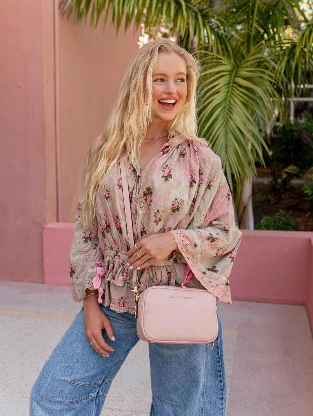 Woman in floral blouse and jeans holding a pink crossbody bag outdoors.
