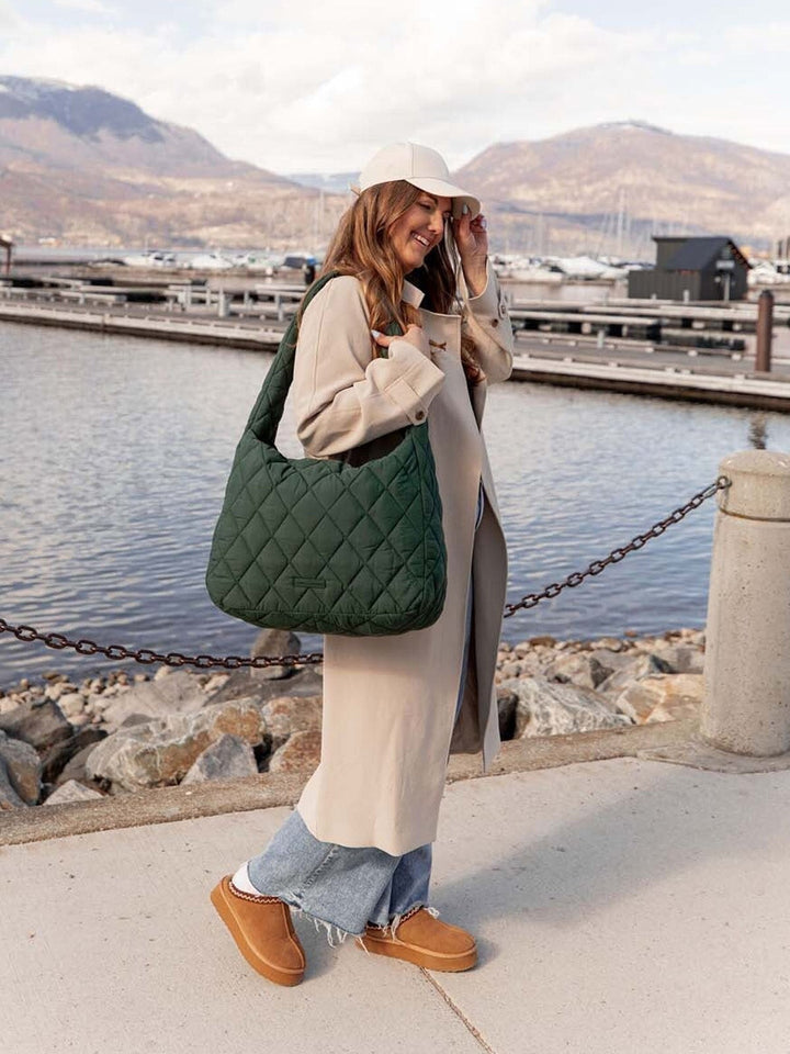 Woman holding a green quilted bag by a waterfront with mountains in the background