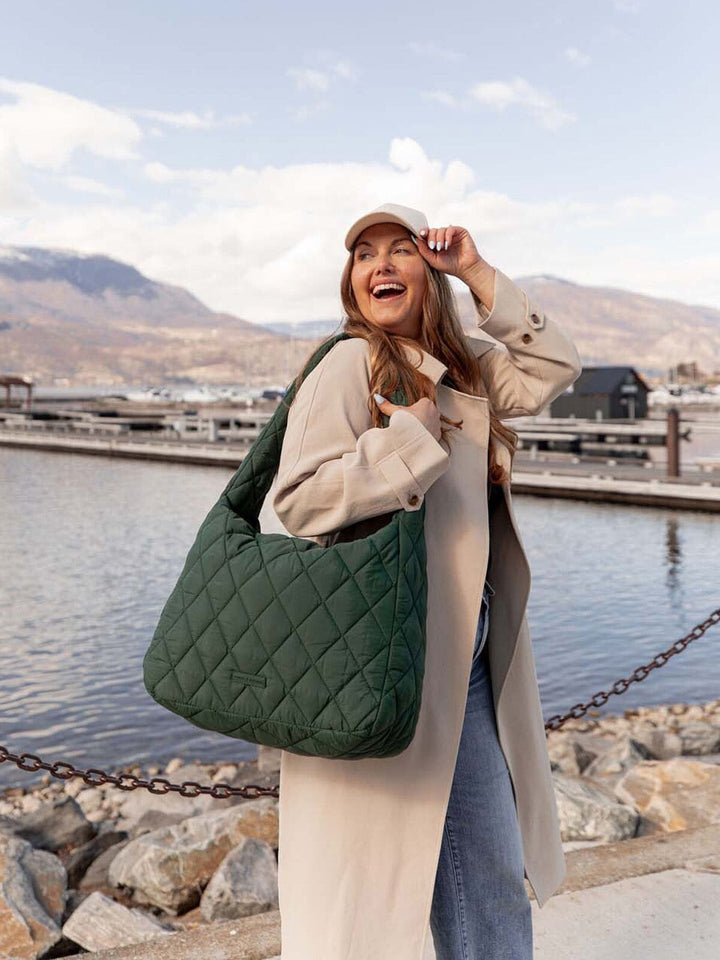 Woman holding a green quilted bag by a waterfront with mountains in the background