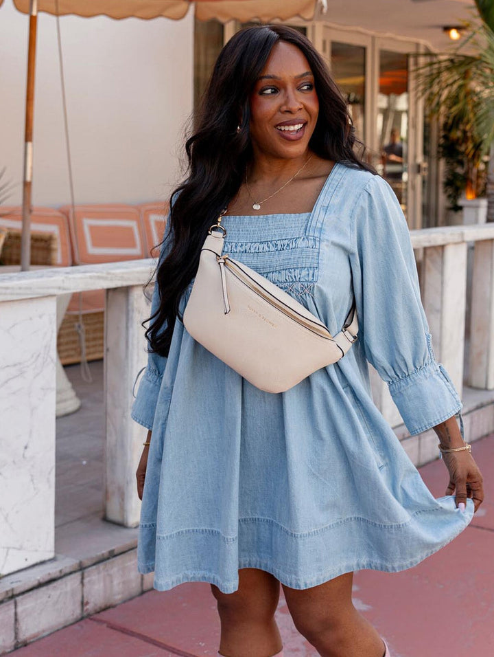 Woman in a light blue dress holding a cream crossbody bag outdoors.