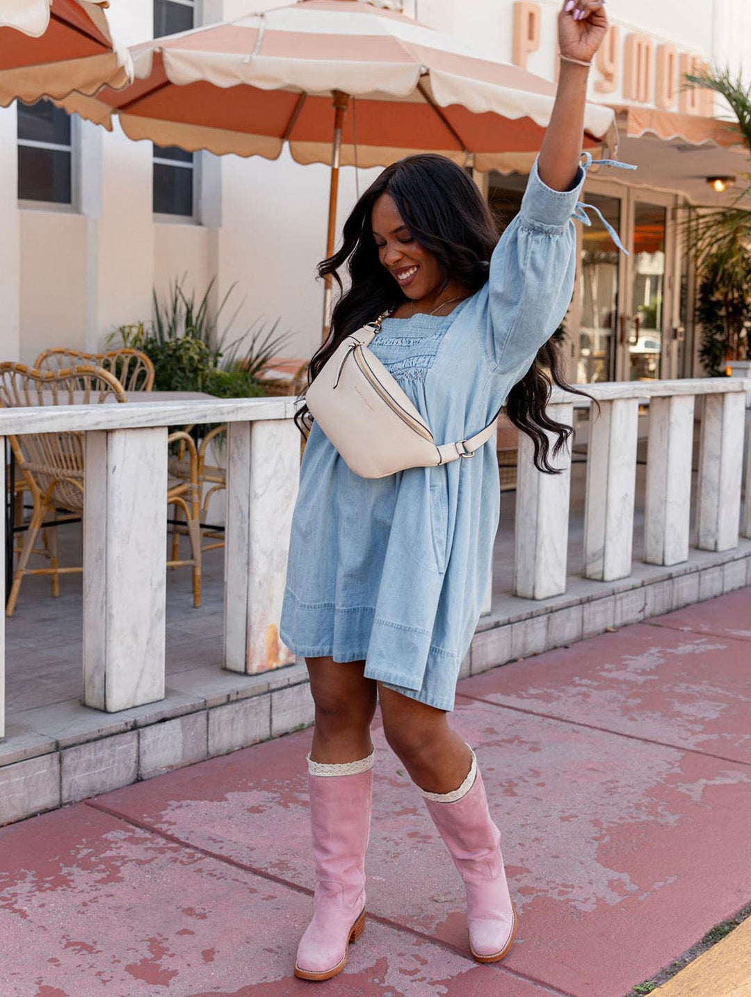 Woman in a denim dress, pink boots  and a cream crossbody bag standing outdoors with tables and chairs in the background.