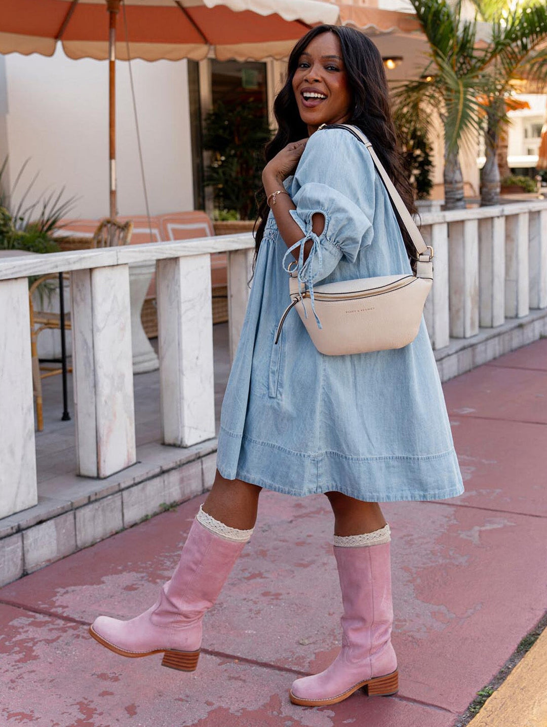Woman in a denim dress and pink boots standing outdoors with a cream handbag.