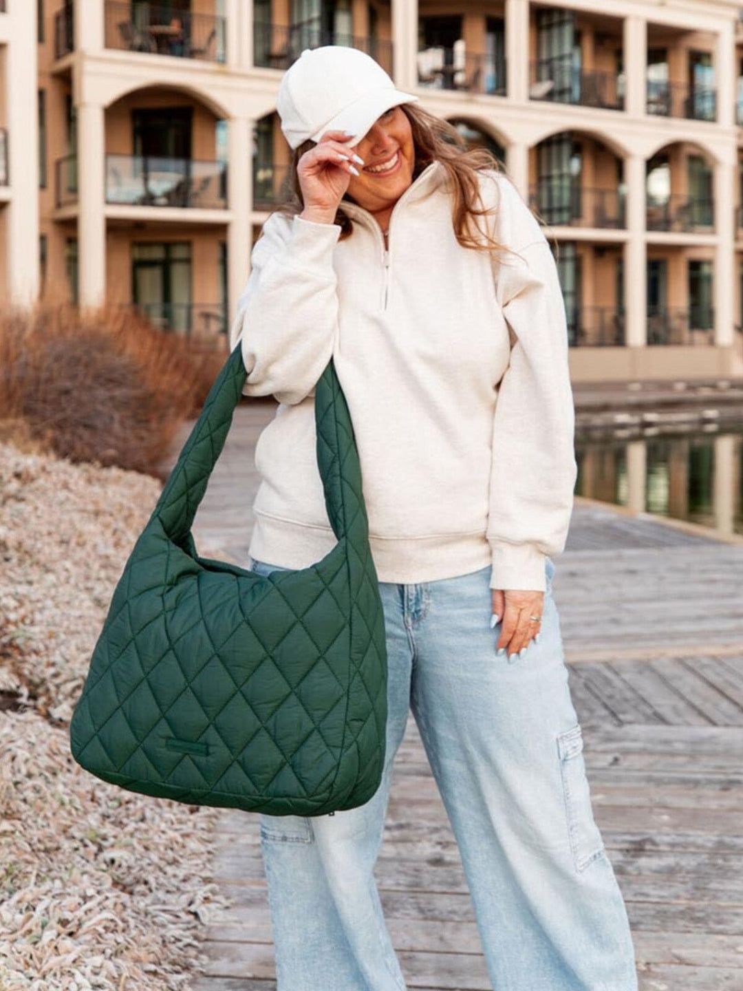 Person wearing a white hoodie and light blue jeans, holding a green quilted bag outdoors.