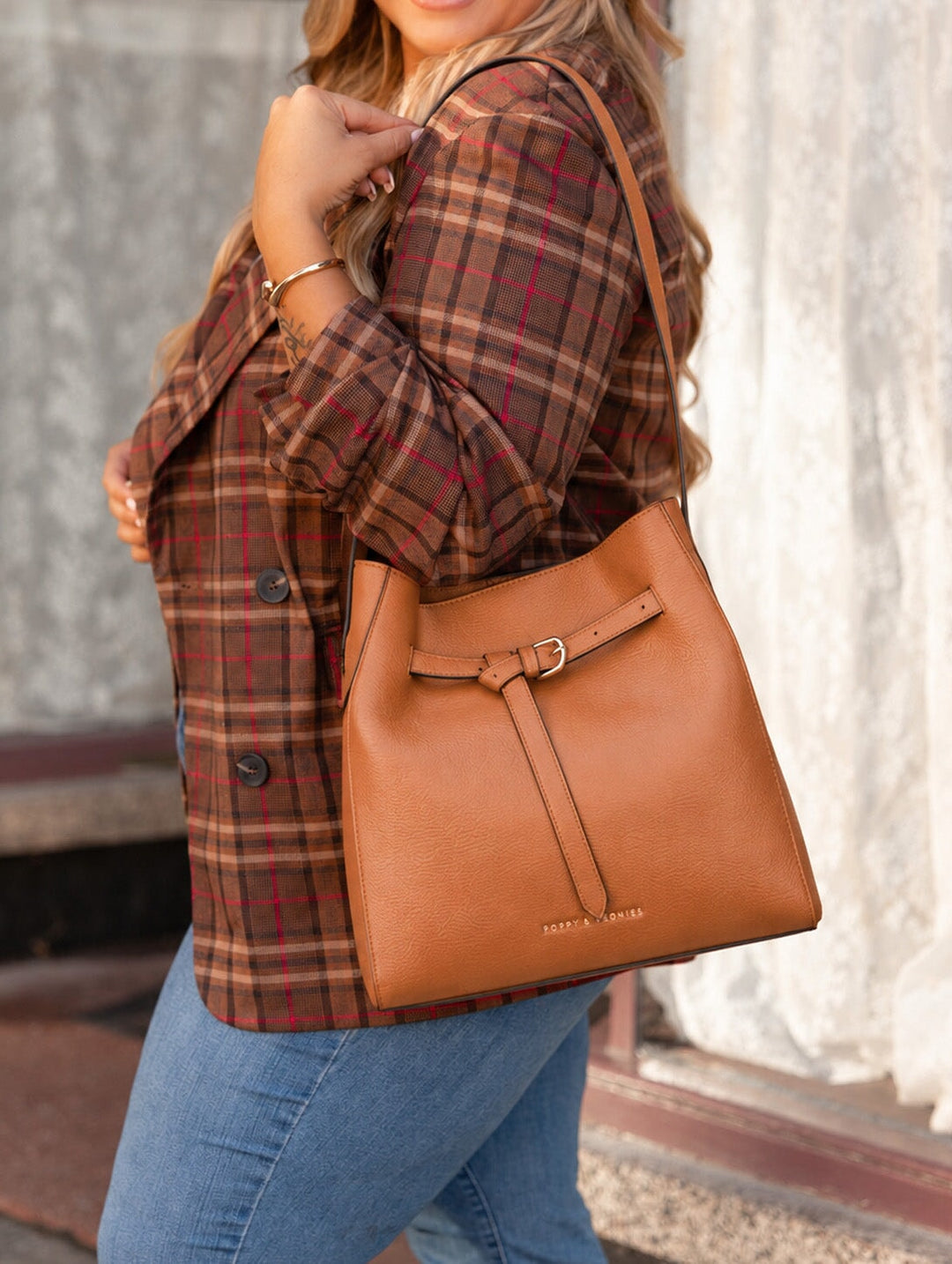 Poppy and Peonies The Knot Shoulder Bag Cognac worn over the shoulder by a woman in a brown plaid blazer and blue jeans, highlighting the bag's structured shape, front buckle detail, and long shoulder strap.