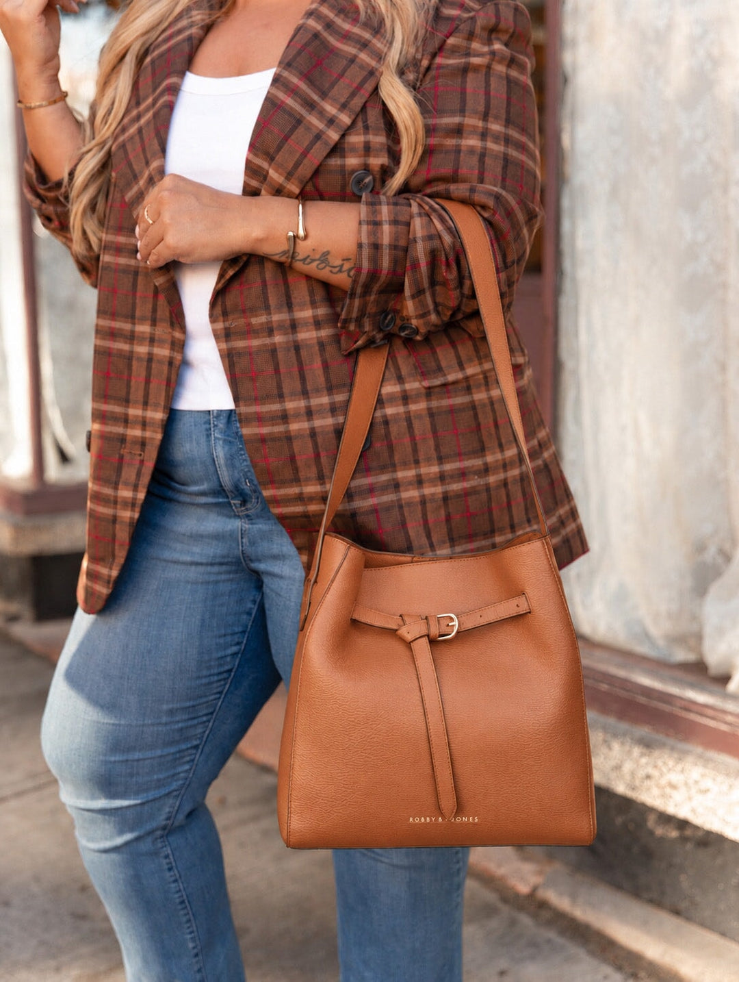 Poppy and Peonies The Knot Shoulder Bag Cognac shown in a close-up lifestyle shot, hanging from a woman's shoulder and highlighting its pebbled texture, adjustable strap, gold-tone buckle, and front strap detail.