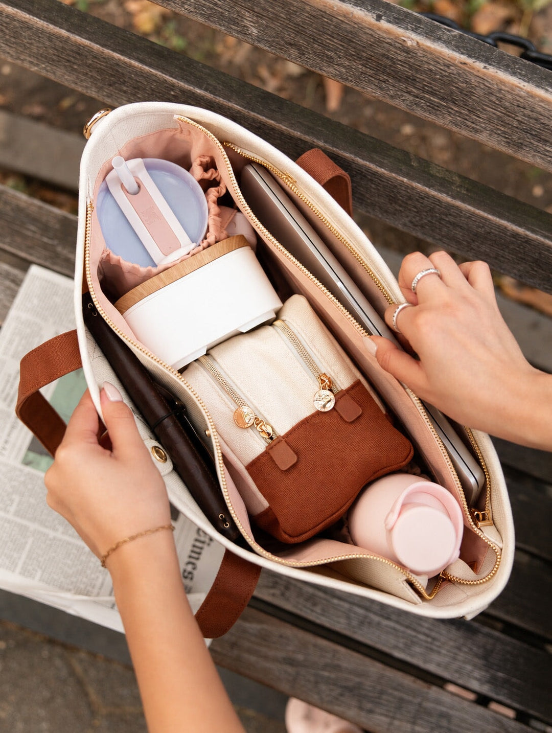 Poppy and Peonies The Signature Tote Chestnut featuring a top-down view of the open tote with blush pink interior lining, multiple organizational compartments, gold-tone zippers, and neatly arranged everyday essentials including a tumbler, cosmetic pouch, water bottle, and tablet, resting on a wooden bench.
